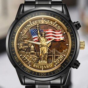 May include: A black wristwatch with a gold-toned face featuring the Statue of Liberty, an American flag, and the text "UNITED STATES", "1776", "RICHARD", and "2026". The watch has a black band and gold accents.