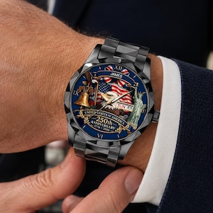 May include: A black metal wristwatch with a blue dial featuring a graphic of the American flag, a bald eagle, the Liberty Bell, and the Statue of Liberty. The dial includes the text JAMES, UNITED STATES OF AMERICA, 250th ANNIVERSARY, 1776-2026.