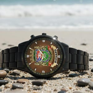 May include: Black wristwatch with a brown face featuring a coat of arms design and the name "RICHARD" on a banner. The watch is set on a sandy beach with small pebbles.
