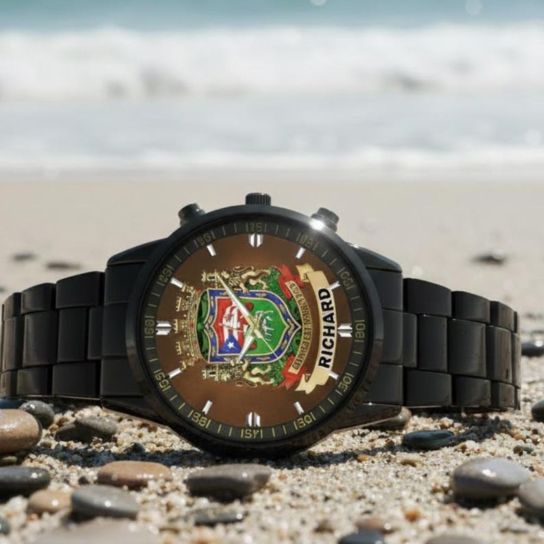 May include: Black wristwatch with a brown face featuring a coat of arms design and the name "RICHARD" on a banner. The watch is set on a sandy beach with small pebbles.