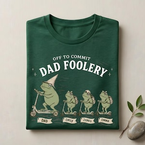 May include: A dark green t-shirt with the slogan "OFF TO COMMIT DAD FOOLERY." The design shows four cartoon frogs on scooters; the first frog wears a wizard's hat. Names "DAD," "EMILY," "JAMES," and "EMMA" are below each frog.