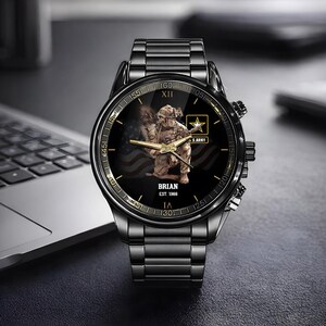 Puede incluir: Reloj de pulsera negro con una correa de metal negro. La esfera del reloj presenta un soldado arrodillado frente a una bandera estadounidense, el texto "BRIAN EST. 1988" y el logotipo del ej&eacute;rcito de EE. UU. El reloj tiene detalles dorados.
