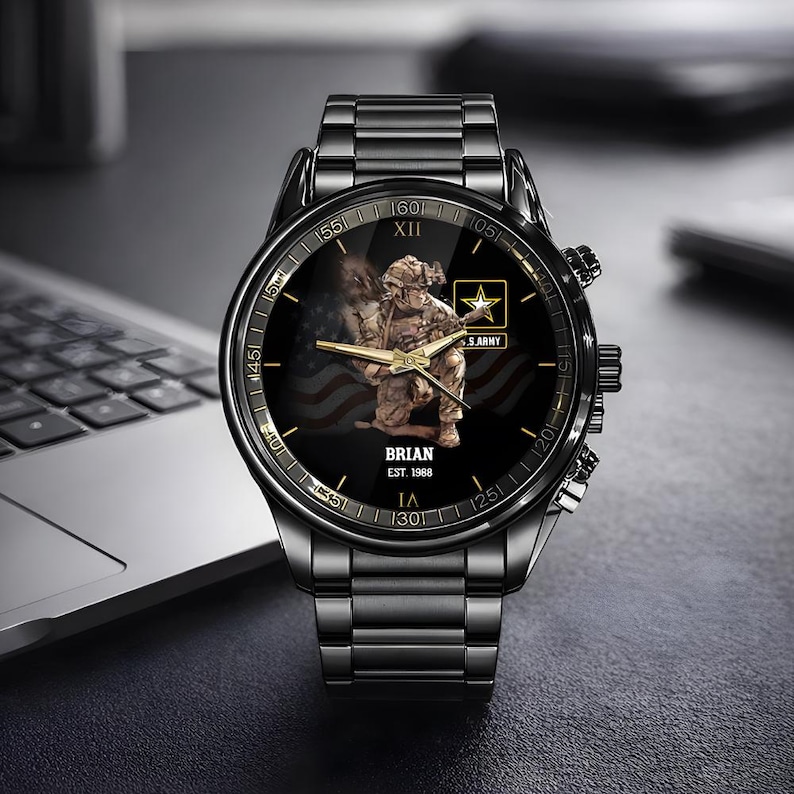 Puede incluir: Reloj de pulsera negro con una correa de metal negro. La esfera del reloj presenta un soldado arrodillado frente a una bandera estadounidense, el texto "BRIAN EST. 1988" y el logotipo del ej&eacute;rcito de EE. UU. El reloj tiene detalles dorados.