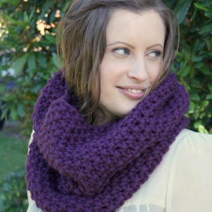 May include: A close-up of a purple infinity scarf. The scarf is made of a thick, chunky knit material. The color is a deep, rich purple. The scarf is wrapped around the neck.