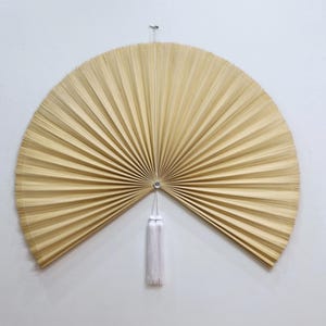 May include: A large, semi-circular wall fan crafted from light beige, thin, radiating slats of natural material. The fan features a central silver-toned circular fastener and a long, white, multi-stranded decorative tassel hanging from the center.