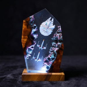May include: A handcrafted resin sculpture featuring a space scene with a Millennium Falcon and X-wing fighters. The artwork is set within a clear resin block, with a wooden base and a light at the bottom. The sculpture has a dark background and a detailed, colorful design.