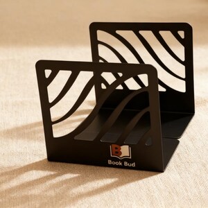May include: Black metal bookends with a wave-like cut-out design. The bookends are angled and feature the text "Book Bud" with a logo. The bookends are on a neutral-colored surface.