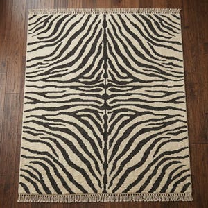 May include: A rectangular area rug featuring a high-contrast zebra stripe pattern in black and cream. The rug has a symmetrical design radiating from the centre and is finished with short, knotted fringe detailing along the top and bottom edges.