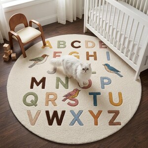 May include: A round, cream-coloured area rug featuring the alphabet in various muted colours, including yellow, green, brown, pink, and blue. The rug is decorated with embroidered illustrations of three small birds positioned among the letters.