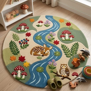 Op de afbeelding: Een rond vloerkleed met een bosscène, een kronkelende blauwe rivier, een slapend reekalfje, een uil op een tak en diverse bosdieren. Het ontwerp bevat groene heuvels, rood-witte paddenstoelen, kleurrijke herfstbladeren en paarse bloemen.