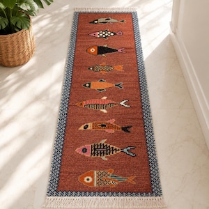 May include: A rectangular, rust-colored runner rug with a repeating pattern of stylized fish in various colors. The rug has a blue and white border and a fringed edge. The fish designs include black, pink, orange, and green.