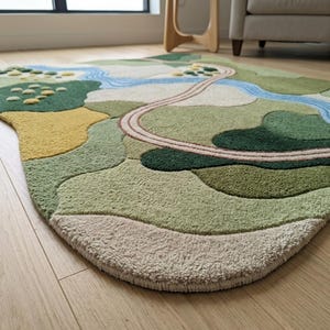 Op de afbeelding: Een getuft vloerkleed met een abstract landschapsontwerp in diverse texturen en kleuren, waaronder tinten groen, cr&egrave;me, geel en blauw. Het patroon toont kronkelende paden, een stromende rivier en afgeronde vormen die doen denken aan heuvels.