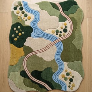 Op de afbeelding: Een vloerkleed met een onregelmatige, golvende vorm en een landschapsontwerp. Het patroon bevat een kronkelende lichtblauwe rivier, een gebogen beige pad en abstracte vlakken in donkergroen, olijfgroen, cr&egrave;me en gele cirkelvormige accenten.