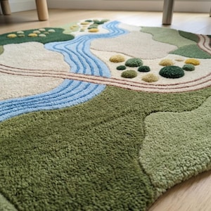 Op de afbeelding: Een getuft karpet met een landschapsontwerp met een kronkelende blauwe rivier, beige paden en stukken groen en geel getextureerd terrein. Het kleed heeft een hoogpolig oppervlak met verschillende tinten groen, cr&egrave;me en blauw, wat een topografisch kaarteffect cre&euml;ert met verhoogde elementen over het gehele oppervlak.