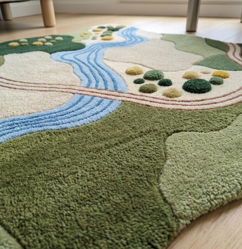 Op de afbeelding: Een getuft karpet met een landschapsontwerp met een kronkelende blauwe rivier, beige paden en stukken groen en geel getextureerd terrein. Het kleed heeft een hoogpolig oppervlak met verschillende tinten groen, cr&egrave;me en blauw, wat een topografisch kaarteffect cre&euml;ert met verhoogde elementen over het gehele oppervlak.