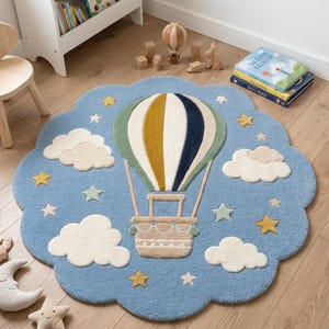 Op de afbeelding: Een rond vloerkleed met geschulpte rand in lichtblauw, voorzien van een centrale heteluchtballon met verticale strepen in mosterdgeel, wit en donkerblauw. Het ontwerp bevat witte getufte wolken en verspreide gele, witte en lichtgroene sterren.