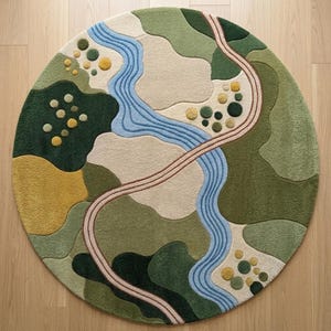 Op de afbeelding: Een rond vloerkleed met een getuft landschapsontwerp met een kronkelende blauwe rivier, een gebogen beige pad en abstracte vlakken in donkergroen, olijfgroen, lichtgroen en mosterdgeel. Het kleed bevat kleine, cirkelvormige getufte accenten.