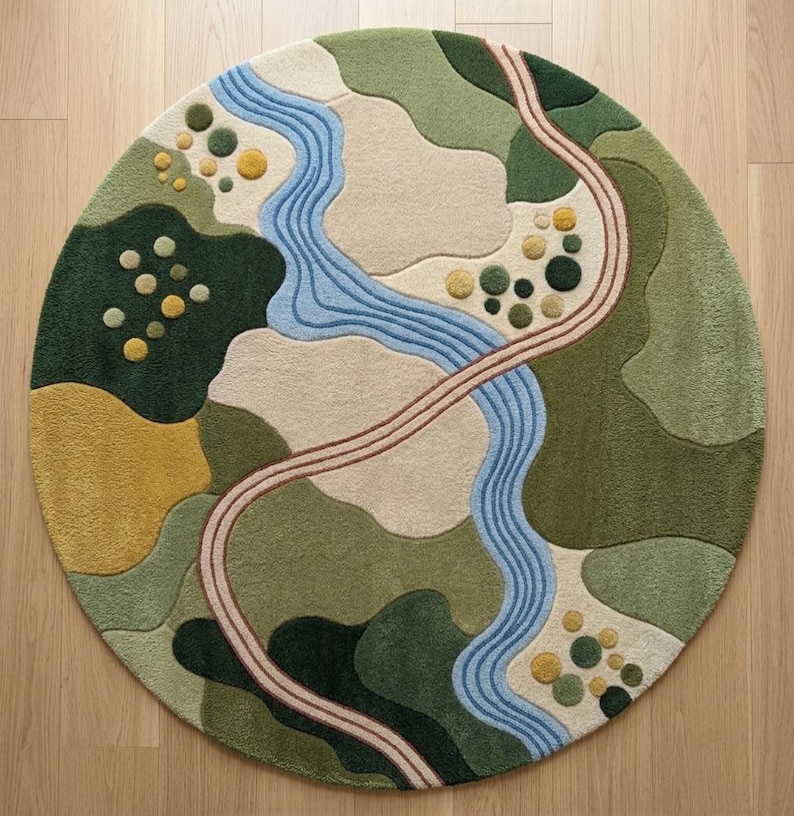 Op de afbeelding: Een rond vloerkleed met een getuft landschapsontwerp met een kronkelende blauwe rivier, een gebogen beige pad en abstracte vlakken in donkergroen, olijfgroen, lichtgroen en mosterdgeel. Het kleed bevat kleine, cirkelvormige getufte accenten.