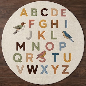 May include: A circular, off-white area rug featuring the alphabet in various muted colours including yellow, green, brown, pink, and blue. The design includes three small bird illustrations interspersed among the letters A through Z in a grid layout.