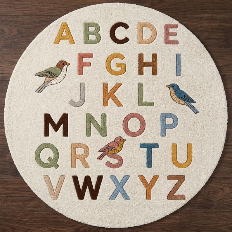 May include: A circular, off-white area rug featuring the alphabet in various muted colours including yellow, green, brown, pink, and blue. The design includes three small bird illustrations interspersed among the letters A through Z in a grid layout.