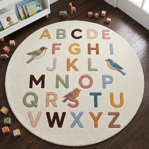 May include: A round, cream-coloured area rug featuring the alphabet in raised, multi-coloured letters. The design includes illustrations of three birds in shades of green, blue, and pink, interspersed among the letters A through Z in a grid.