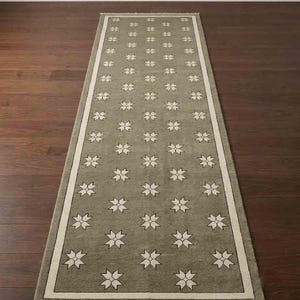 May include: A long rectangular runner rug featuring a muted olive green base color with a repeating pattern of white four-pointed star motifs. The rug is framed by a solid white border and finished with short white fringe along both narrow ends.