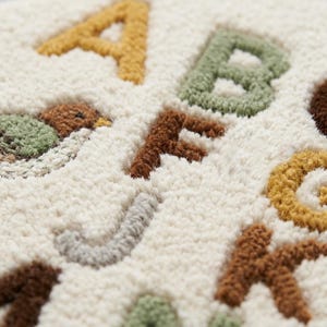May include: A cream-coloured tufted textile featuring raised, embroidered letters of the alphabet in various colours including mustard yellow, sage green, and brown. The surface has a thick, textured pile with a visible bird illustration.