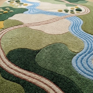 Op de afbeelding: Een getuft tapijt met een landschapsontwerp in tinten groen, beige en blauw. Het patroon toont een kronkelende blauwe rivier, een bruin pad en getextureerde cirkelvormige figuren die bomen of struiken voorstellen in een heuvelachtig terrein.