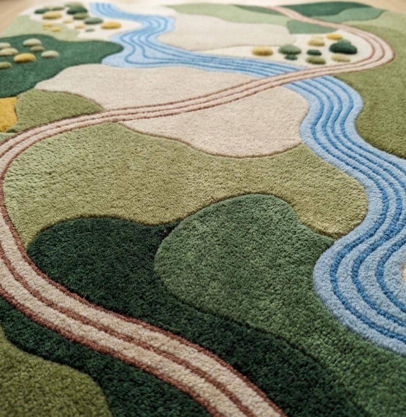 Op de afbeelding: Een getuft tapijt met een landschapsontwerp in tinten groen, beige en blauw. Het patroon toont een kronkelende blauwe rivier, een bruin pad en getextureerde cirkelvormige figuren die bomen of struiken voorstellen in een heuvelachtig terrein.