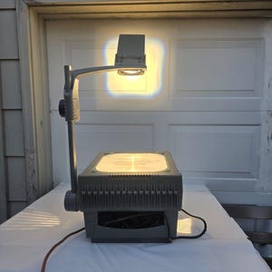 May include: A gray overhead projector featuring a rectangular base with a glass stage, a vertical support arm, and an adjustable projection head. The device is powered on, emitting a bright, warm yellow light from the lens onto the wall.