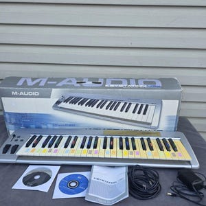 May include: A silver M-Audio Keystation 49es MIDI keyboard with black and white keys, colorful accents, and a matching box. Includes a user guide, CDs, and cables. The keyboard is approximately 32 inches wide.
