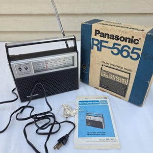Könnte beinhalten: Ein tragbares Panasonic RF-565 FM-AM Radio mit schwarz-silbernem Gehäuse, Teleskopantenne und schwarzem Netzkabel. Das Set enthält den originalen Karton mit Produkttext, einen Ohrhörer und eine Bedienungsanleitung.