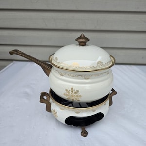 Puede incluir: Una olla para fondue de esmalte color crema con tapa a juego y mango largo, con patrones de copos de nieve dorados y adornos decorativos. La olla descansa sobre un soporte de metal color crema y dorado a juego con tres patas.