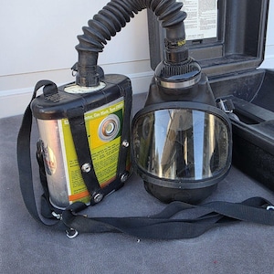 Puede incluir: Una máscara de gas MSA negra con una visera transparente y un tubo respiratorio corrugado. La máscara está conectada a un recipiente cilíndrico con una etiqueta amarilla y negra. Se adjuntan correas negras para usar la máscara.