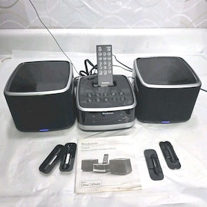 May include: A black Brookstone Stack stereo system with two speakers, a central control unit, and a remote. The speakers have silver trim and a blue light at the base. Accessories and a manual are included.