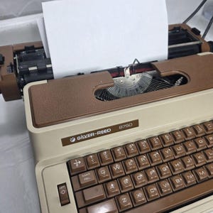 Puede incluir: Una máquina de escribir Silver-Reed 8750 vintage, en marrón y beige. El teclado tiene teclas marrones con letras blancas. Se ha insertado una hoja de papel blanca en blanco. La máquina de escribir está sobre una superficie clara.