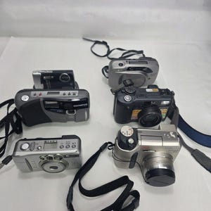 以下が含まれることがあります： シルバー、ブラック、グレーなど、さまざまな色のデジタルカメラ5台のコレクション。各カメラにはレンズ、ボタン、ストラップが付いています。カメラは白い表面に配置されています。