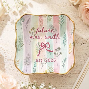 Peut inclure: Un vide-poches carré à rayures pastel et bord doré, orné d'illustrations florales et de feuilles. L'inscription "future mrs. smith est. 2026" est présente dans une écriture délicate, avec un nœud. L'ensemble est mis en valeur par un éclairage doux et des éléments floraux.