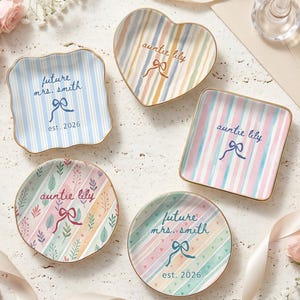 Peut inclure: Assortiment de petits plats décoratifs, ronds, carrés et en forme de cœur, avec des rayures pastel et des motifs floraux. Certains plats portent les inscriptions "future mrs. smith est. 2026" et "auntie lily". Bords dorés.
