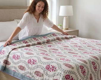 Könnte beinhalten: Eine Tagesdecke mit cremefarbener Basis und einem sich wiederholenden Muster aus rosa Blumenmotiven und graugrünen Ranken. Die Decke ist mit einem einfarbigen hellblauen Rand und einer schmalen blau-weiß gestreiften Kante versehen.