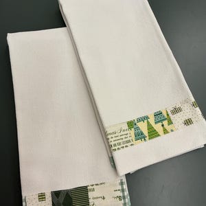 Op de afbeelding: Twee witte theedoeken met wafelstructuur en decoratieve stoffen randen. Eén rand toont groene en gele kerstboompatronen met cadeautjes, terwijl de andere een patchworkontwerp heeft van groene strepen, stippen en cadeau-iconen.