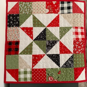 Op de afbeelding: Een vierkante gewatteerde wandhanger met een centraal sterpatroon bestaande uit rode, groene, zwarte en witte stoffen lapjes. De quilt is afgewerkt met een effen rode bies en toont diverse patronen waaronder ruiten, stippen en bloemenprints.