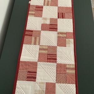 Può includere: Un runner da tavola trapuntato rettangolare con un design patchwork in tessuti rossi e bianchi. Il motivo è composto da quadrati e triangoli alternati in varie stampe a quadretti, righe e pois rossi, rifinito con un bordo in tessuto rosso.