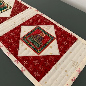 Peut inclure: Un chemin de table matelassé présentant un motif répétitif de tissu rouge à fleurs blanches et de tissu crème avec des contours d'étoiles dorées. Les carrés centraux affichent un motif de cadeau. Une petite étiquette indique Hand made.
