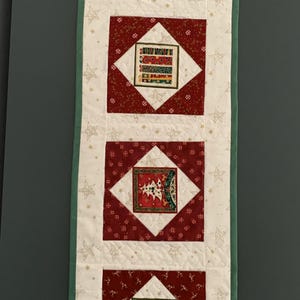 May include: A rectangular quilted table runner featuring a cream-coloured background with gold star patterns. It contains four square panels with dark red borders, each displaying distinct holiday-themed illustrations including trees and stockings.