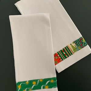 Op de afbeelding: Twee witte theedoeken met wafelstructuur. Eén doek heeft een groene rand met gele sterren en lichtblauwe lintpatronen. De andere doek heeft een veelkleurige rand met rode, groene en witte strepen, kerstbomen en besjes als decoratie.