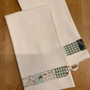 Pode incluir: Dois panos de cozinha brancos com textura de waffle e uma borda de tecido decorativa na parte inferior. A borda inclui padrões de bonecos de neve, um quadrado azul claro com uma figura e o texto "BRR... BLIZZARD candy cane" com um ícone.