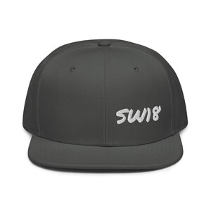 Peut inclure: Casquette de baseball gris foncé avec le texte brodé "SW18" en blanc. La casquette a une visière plate et une couronne structurée, idéale pour un look décontracté.