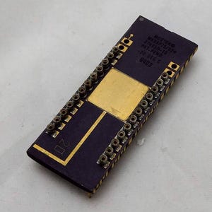 May include: A rectangular electronic component with a dark purple body and gold-coloured accents. The component has numerous pins along the sides and a central gold square. Text on the component includes "MOSTEKO", "MK38P75/92H", and "8403".