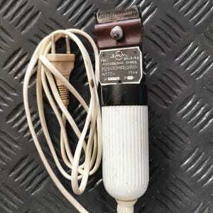 Peut inclure: Tondeuse à cheveux électrique vintage avec une tête en métal marron, une poignée blanche rainurée et un cordon d'alimentation blanc. La tondeuse porte une étiquette avec du texte cyrillique. La prise est de couleur beige clair.